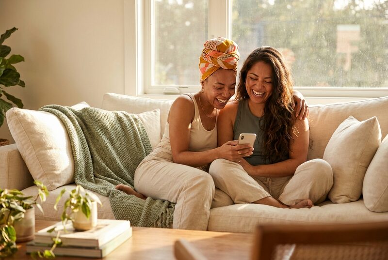 Two friends laugh on a sofa looking at a phone. One wears a bright patterned head wrap.